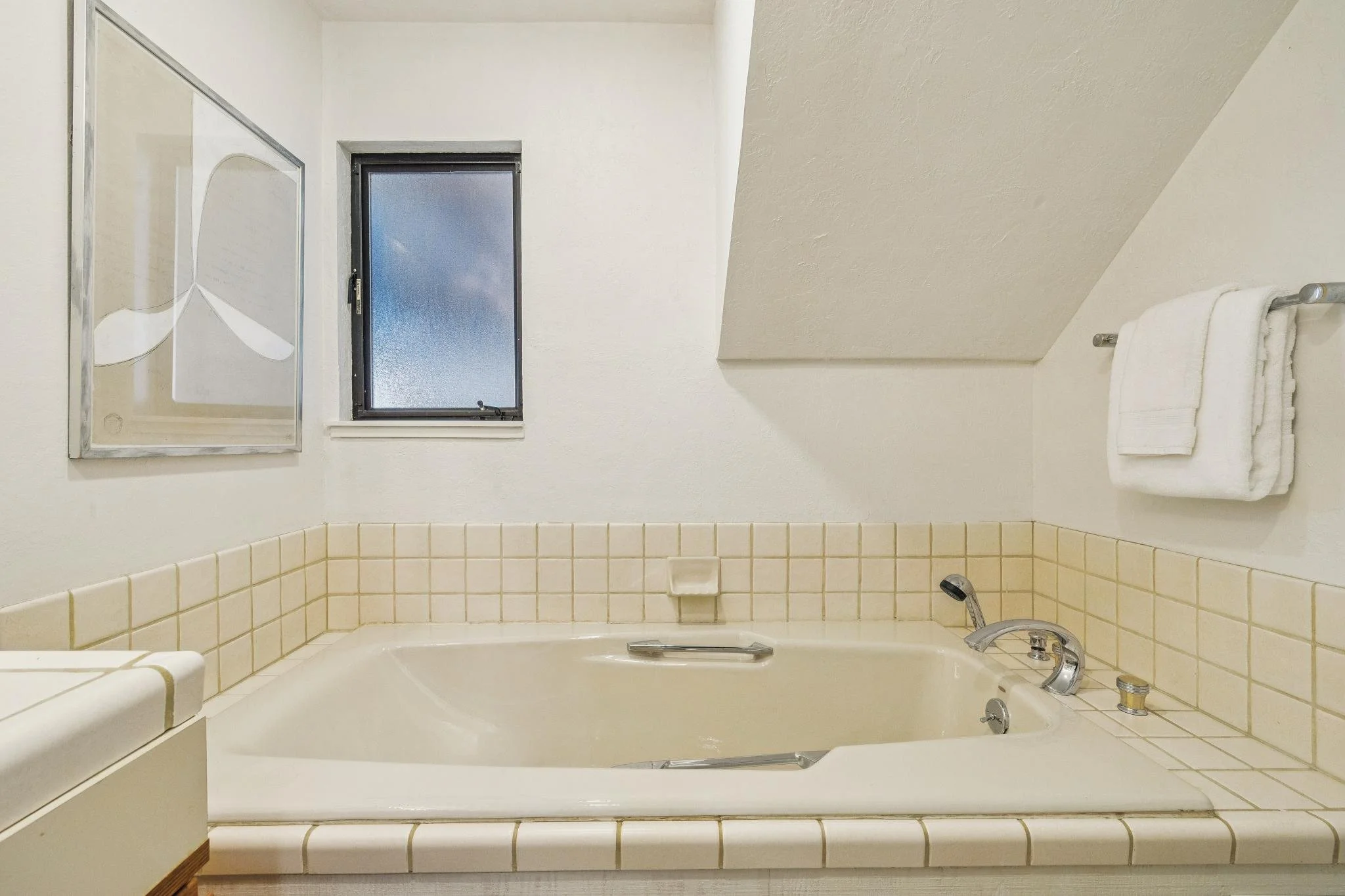 A small bathroom with a bathtub, a tiled surround, a window with frosted glass, a mirror, and a towel rack with white towels.