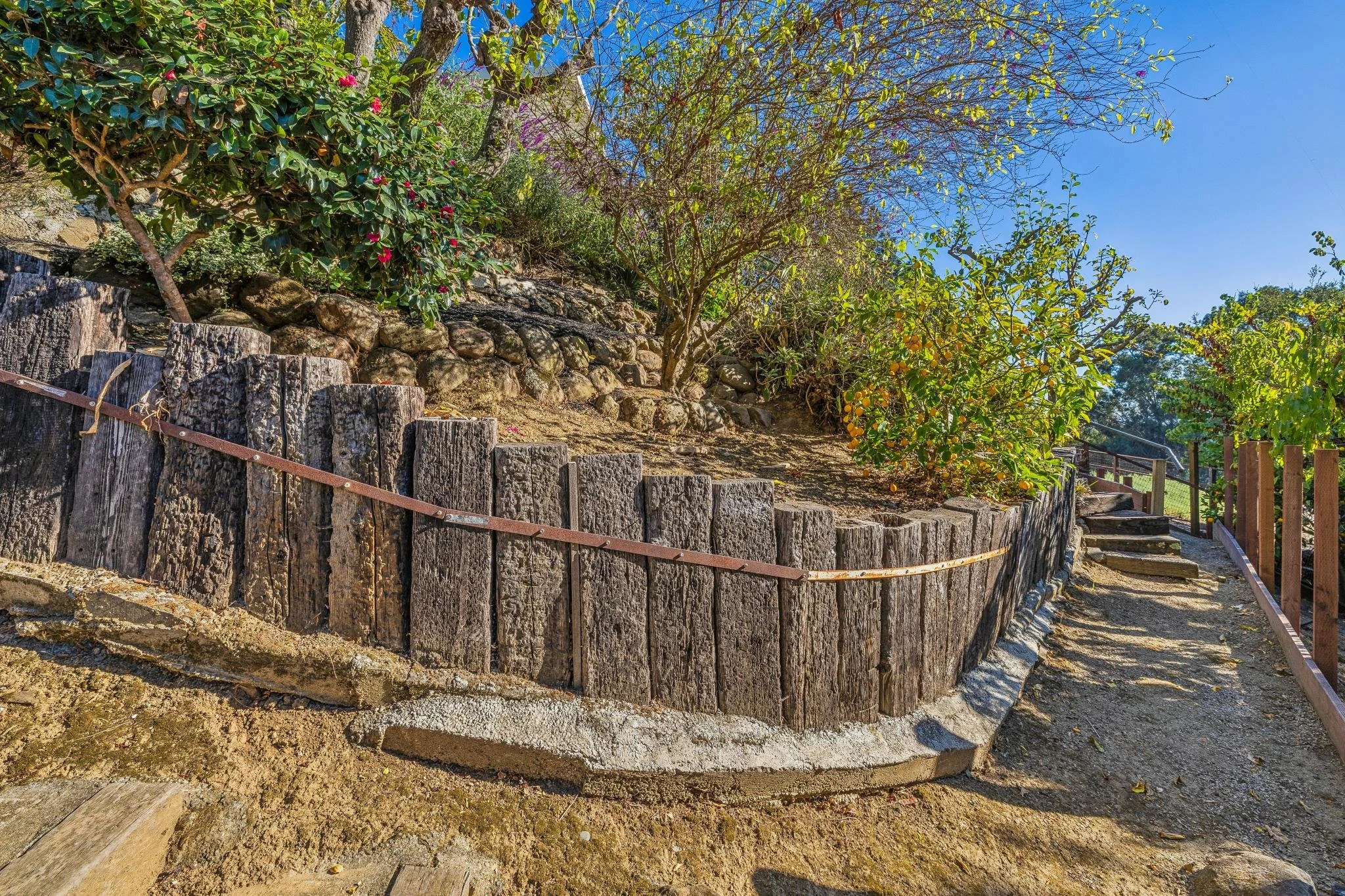 A stone retaining wall with wooden posts and rope, set against a dirt pathway on a slope with trees and shrubs, under a clear blue sky.