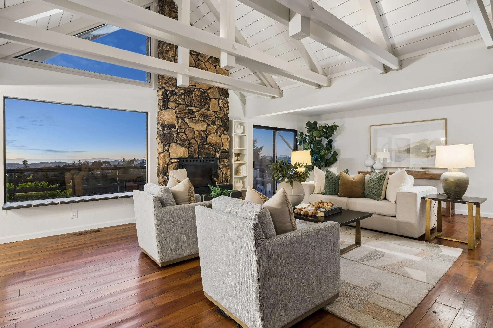 Living room with large windows showing a view of a clear sky, wooden floors, a stone fireplace, white ceiling beams, white walls, a beige sectional sofa with cushions, a gray armchair, a coffee table with decorative objects, a large potted plant, and