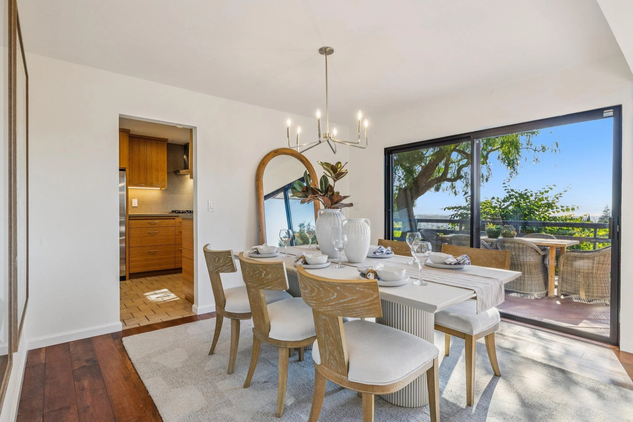 Dining room with a wooden table set with white dishes and glassware, surrounded by wooden chairs with white cushions, large sliding glass door opening to a balcony with outdoor seating and views of trees and blue sky, modern chandelier hanging from c
