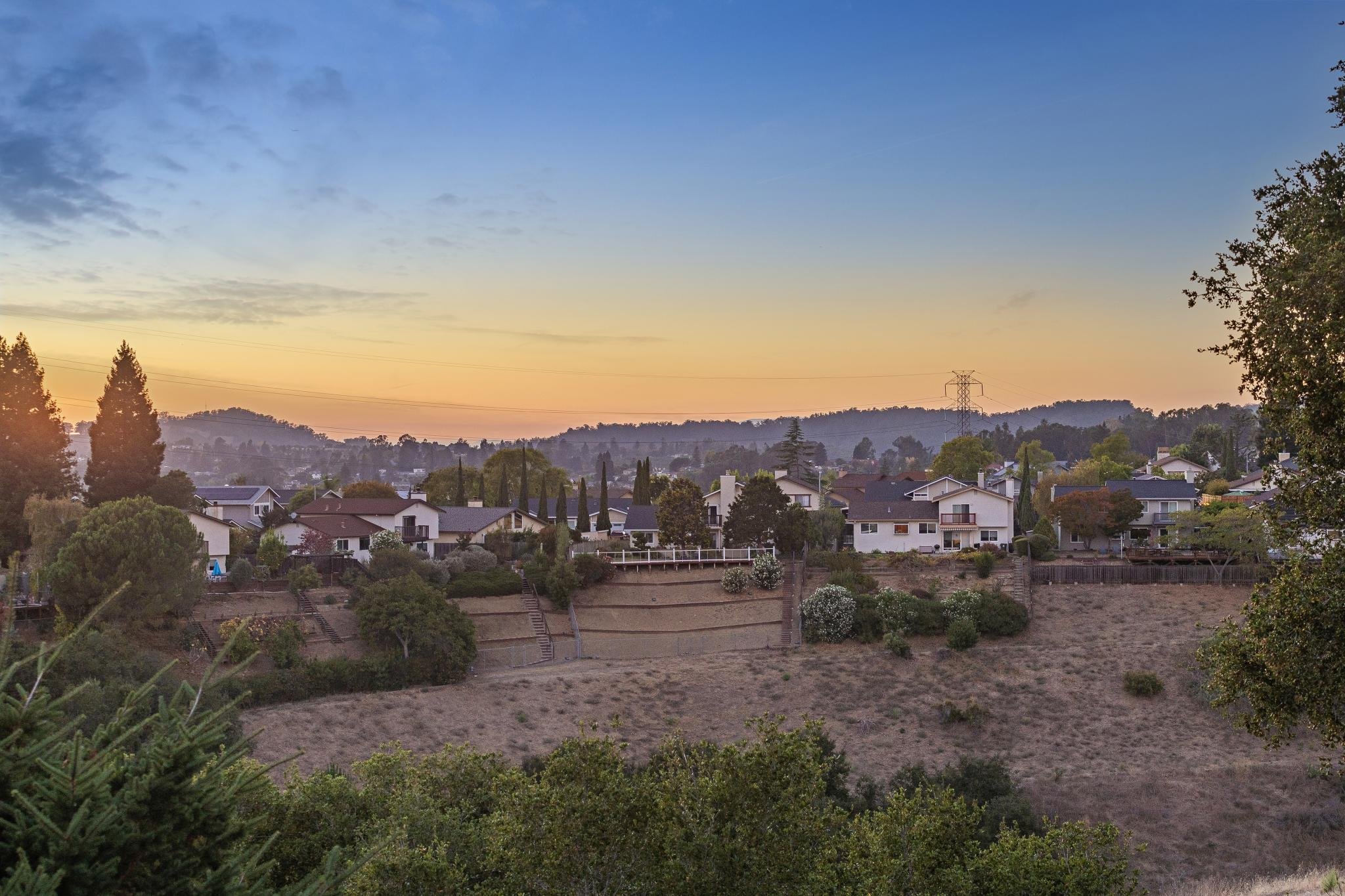 A neighborhood view at sunset with houses, trees, and distant hills under a colorful sky.