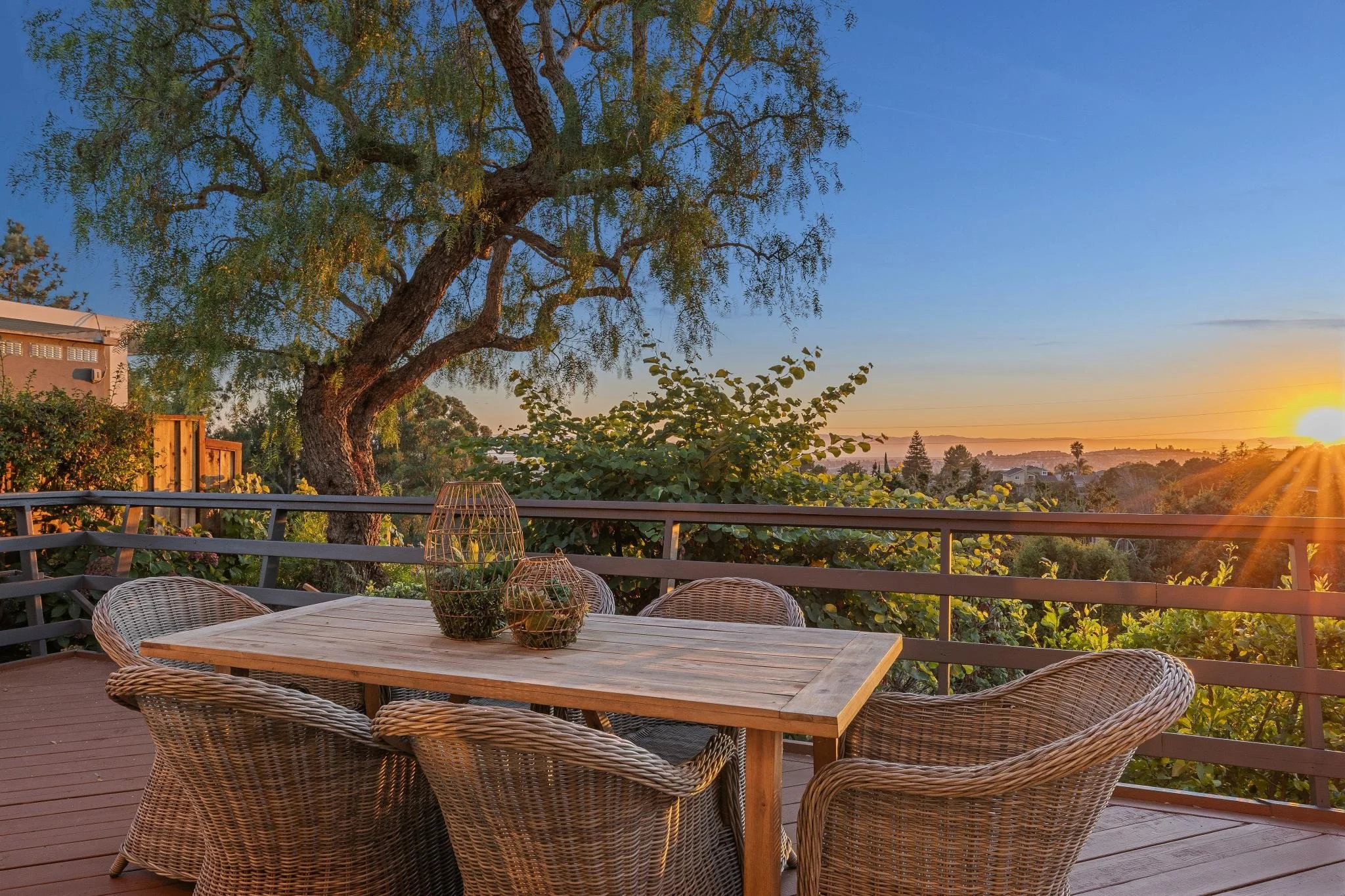 View of a backyard patio at sunset with a wooden table and woven chairs, a large tree, and a scenic landscape with rolling hills and a sunset sky.