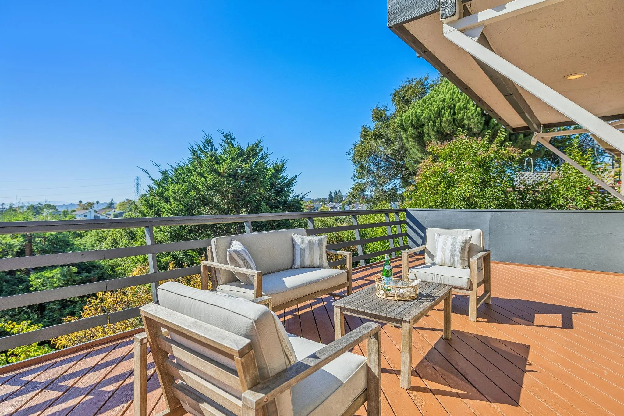 Outdoor balcony with wooden furniture including chairs, a sofa, and a small table with a bottle of sparkling water, overlooking green trees and a clear blue sky.