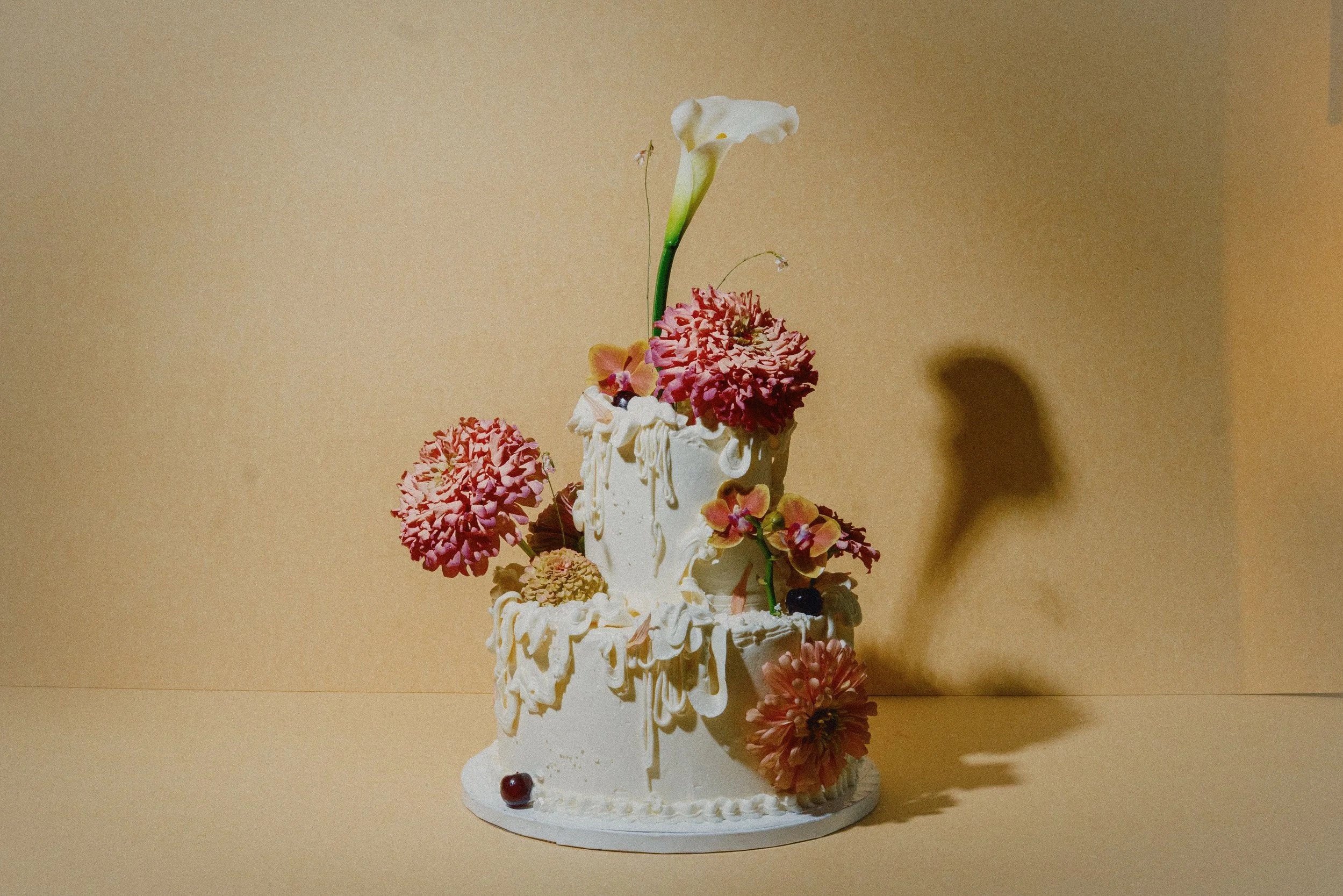 A multi-tiered wedding cake with white cake icing decorated with pink and peach flowers and a single tall white calla lily on top, set against a plain beige background.
