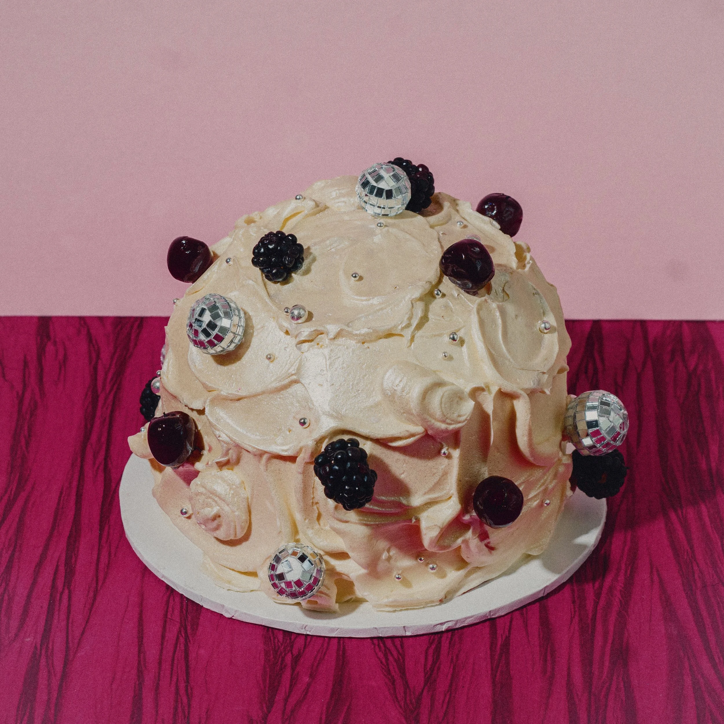 A dome cake decorated with blackberries, cherries, silver pearls, and disco ball ornaments over a pearlescent icing on a white plate with a pink and purple textured background.