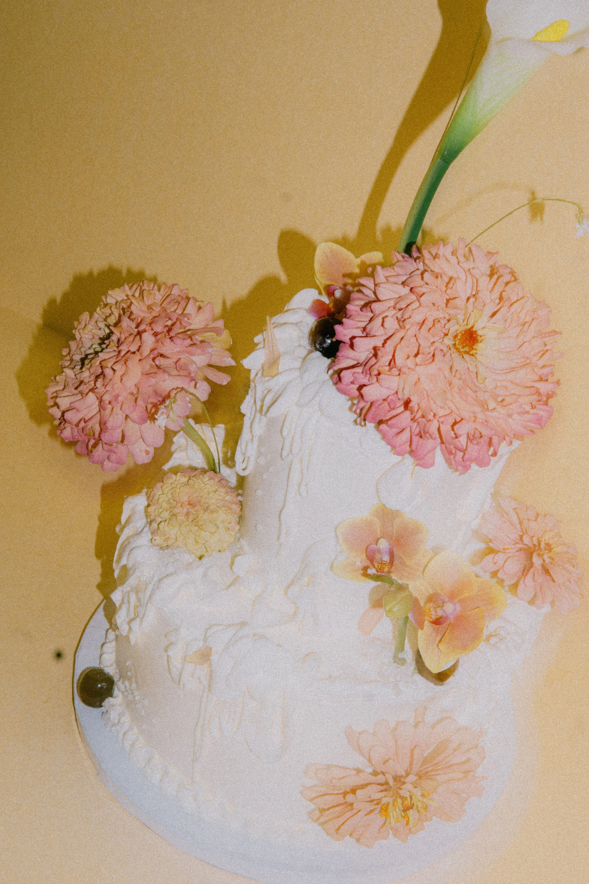A white cake decorated with pink flowers and a single yellow orchid on top. The cake is placed on a reflective surface, and there are blackberries and other small fruits near the flowers.