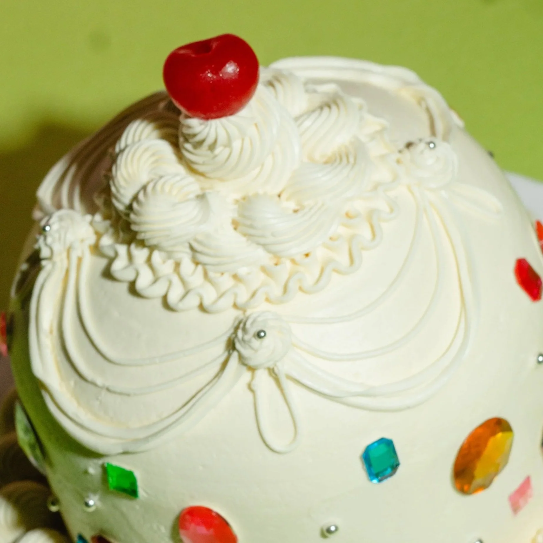 Close-up of a vanilla cake decorated with piped cream, a red cherry on top, and colorful geometric sugar candies around the edge.