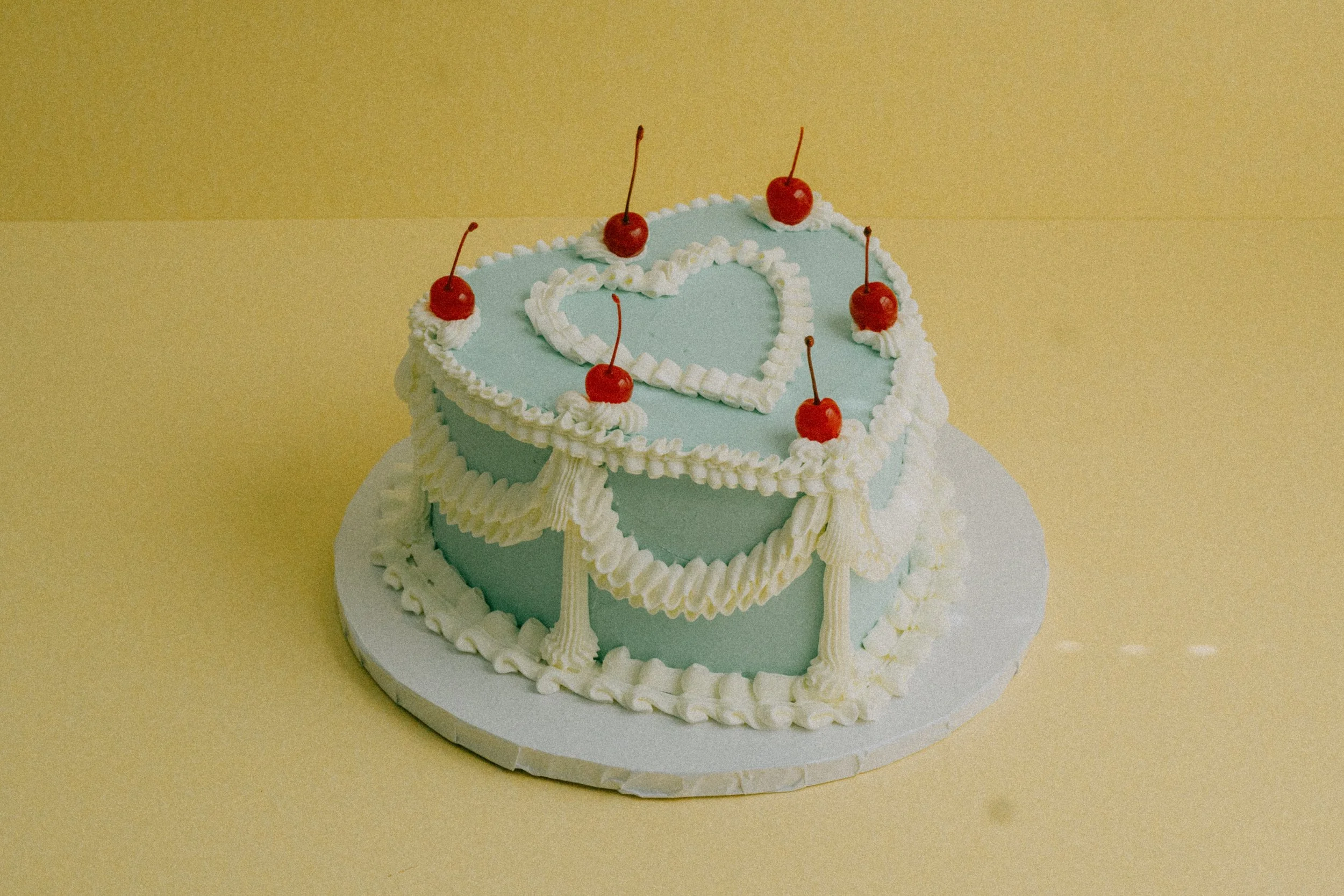 A heart shaped birthday cake with vintage piping and blue buttecream, decorated with white icing trim and a white heart in the center, topped with five red cherries.