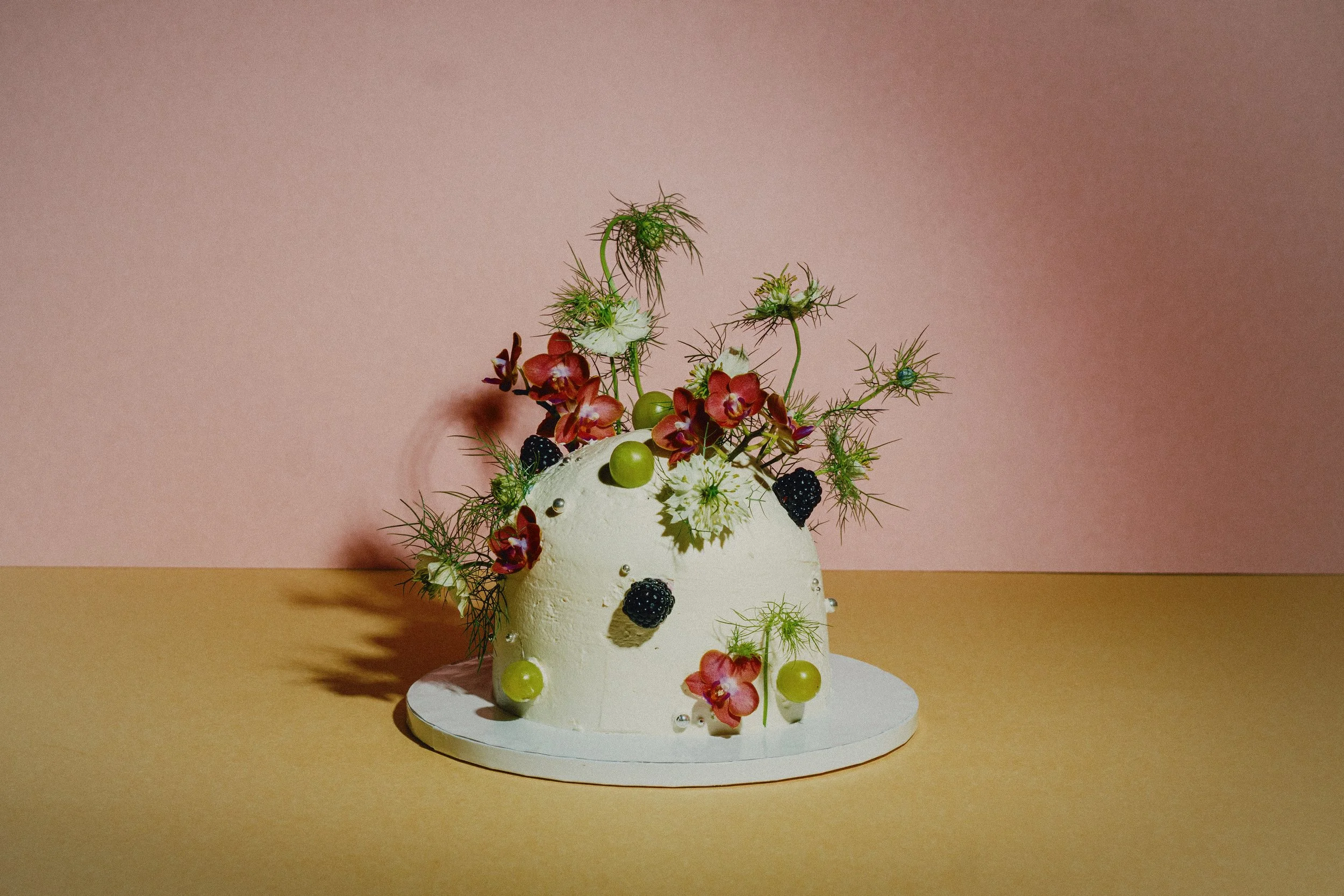 A white cake decorated with blackberries, green grapes, and pink flowers, topped with an arrangement of greenery and white flowers, set on a white doily against a pink background.