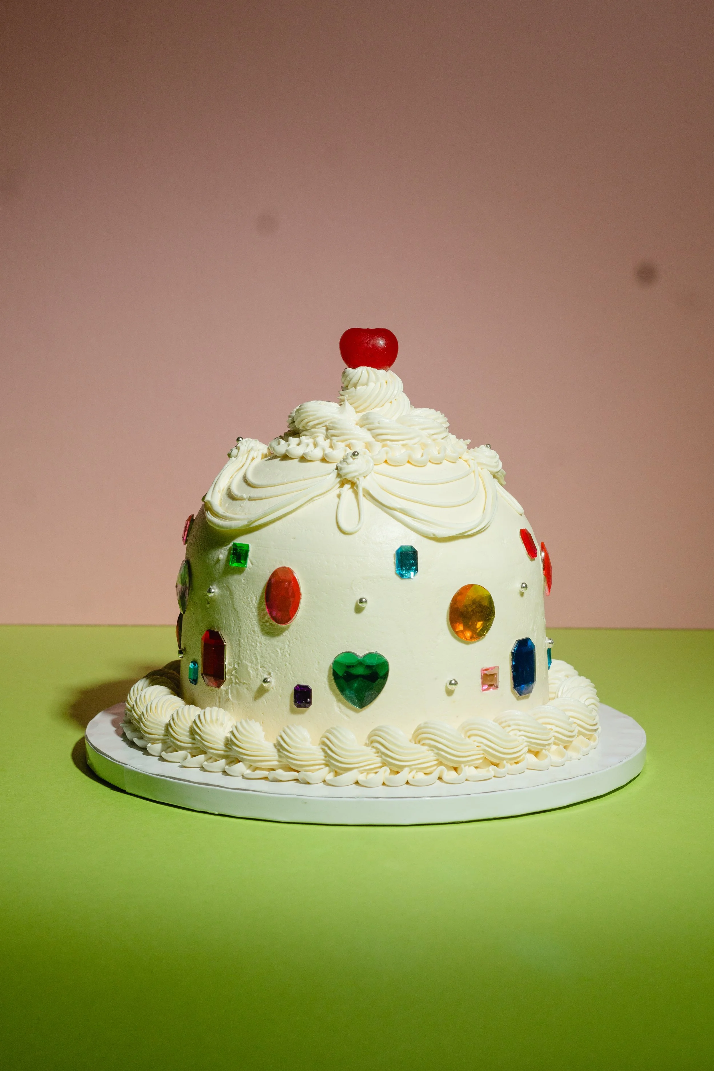 A white birthday cake decorated with colorful gemstones and a cherry on top, with vintage style piping. Perfect for a girls night, bachelorette party or y2k party. Cool Custom cakes