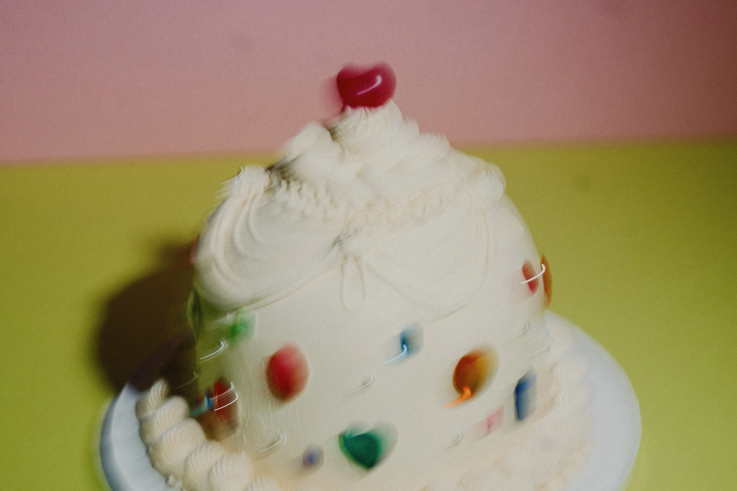 A white birthday cake decorated with colorful round candies and topped with a red heart-shaped decoration, set against a pink and light green background.
