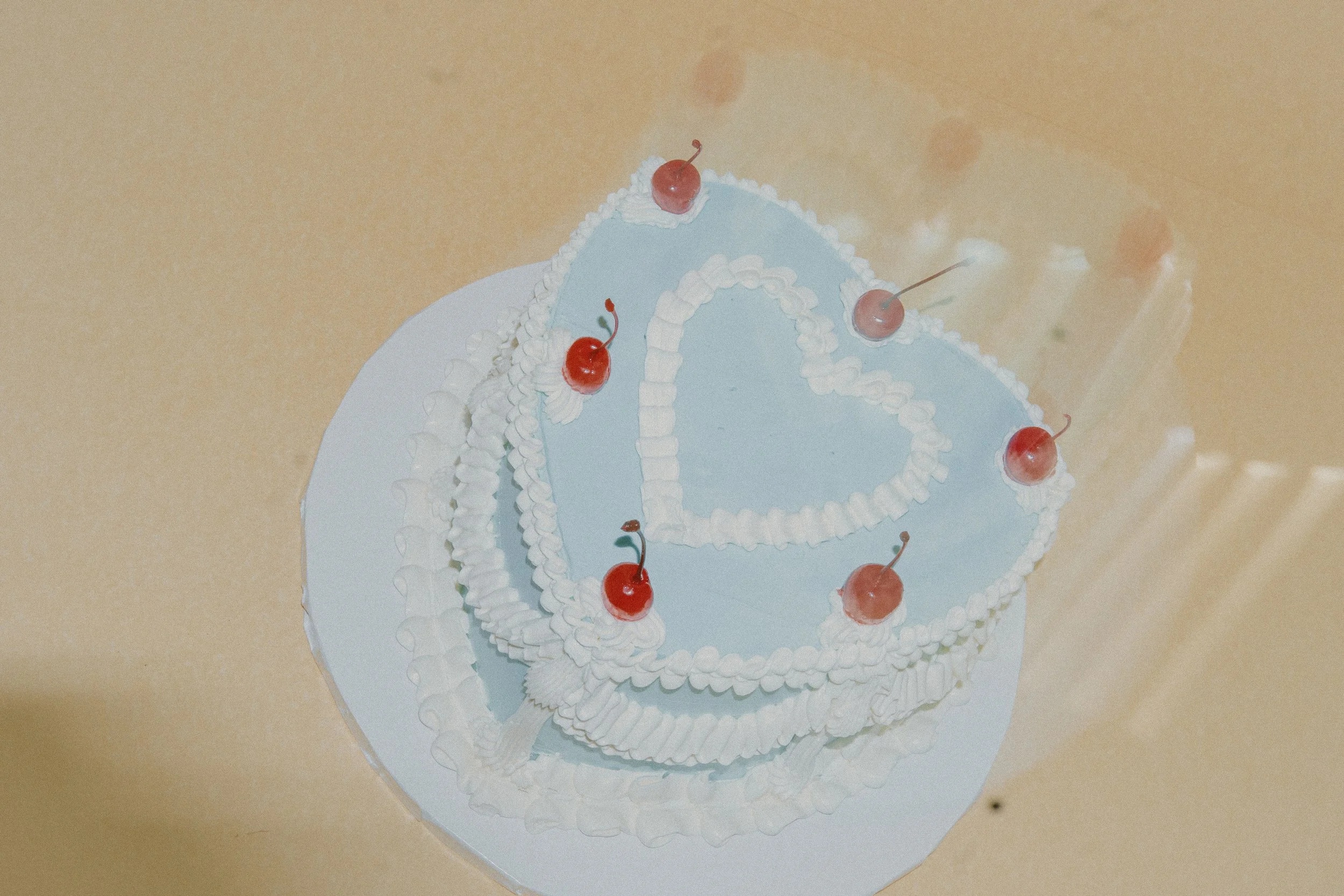 A layered cake with light blue icing, decorated with white frosting details and red cherries on top, on a white doily, placed on a beige surface.