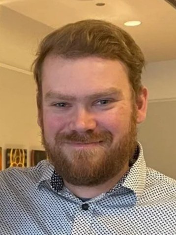 A man with a beard and short brown hair smiling at the camera, wearing a light-colored, patterned collared shirt indoors.