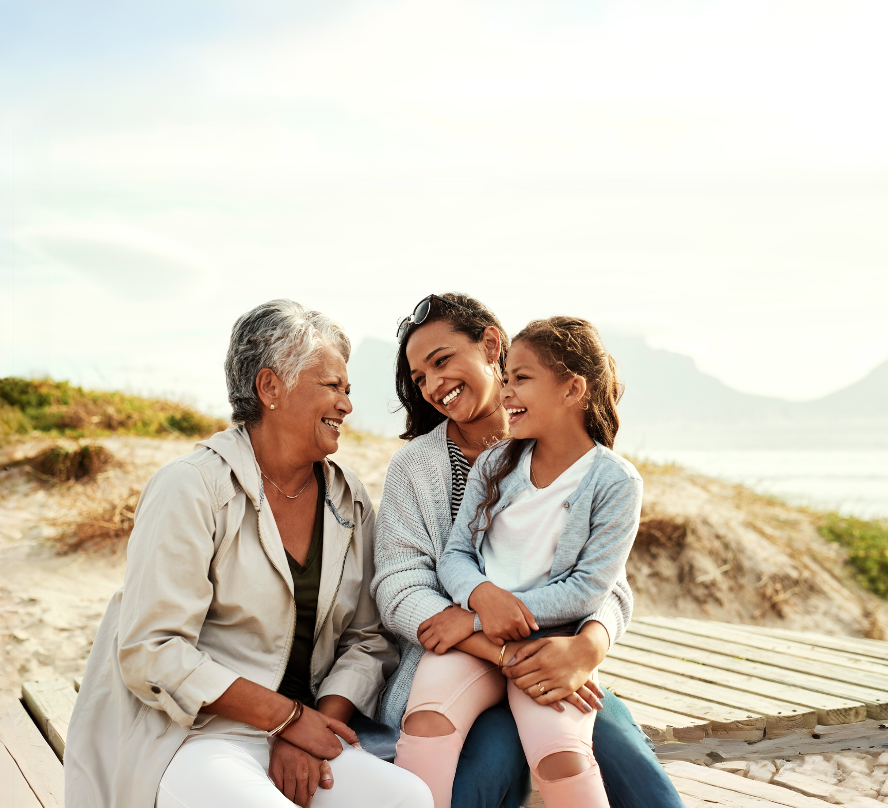 Three women of different generations sitting together on a wooden bench outdoors, smiling and enjoying each other's company on a sunny day at the beach.