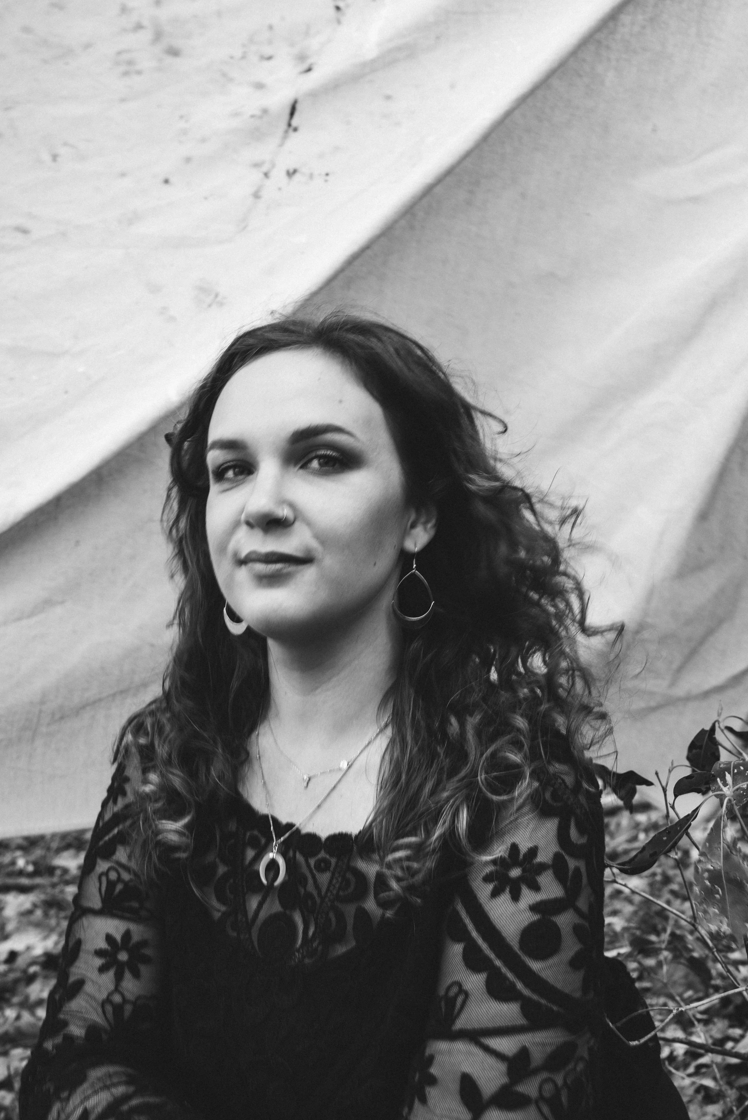 Black and white photo of a woman with curly hair wearing earrings, necklaces, and a floral dress outdoors.