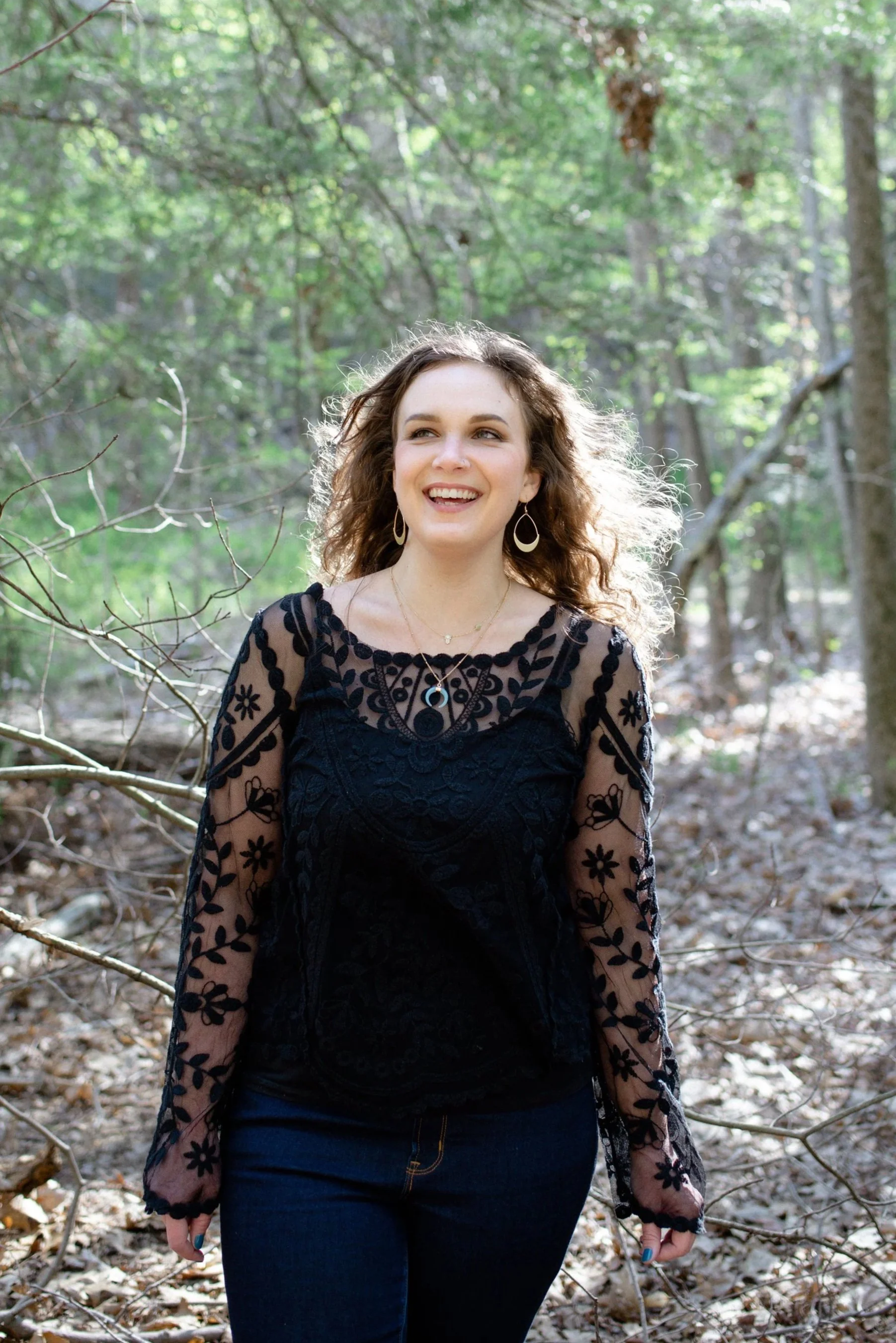 A young woman with brown curly hair smiling in a wooded area, wearing a black lace top and dark jeans.