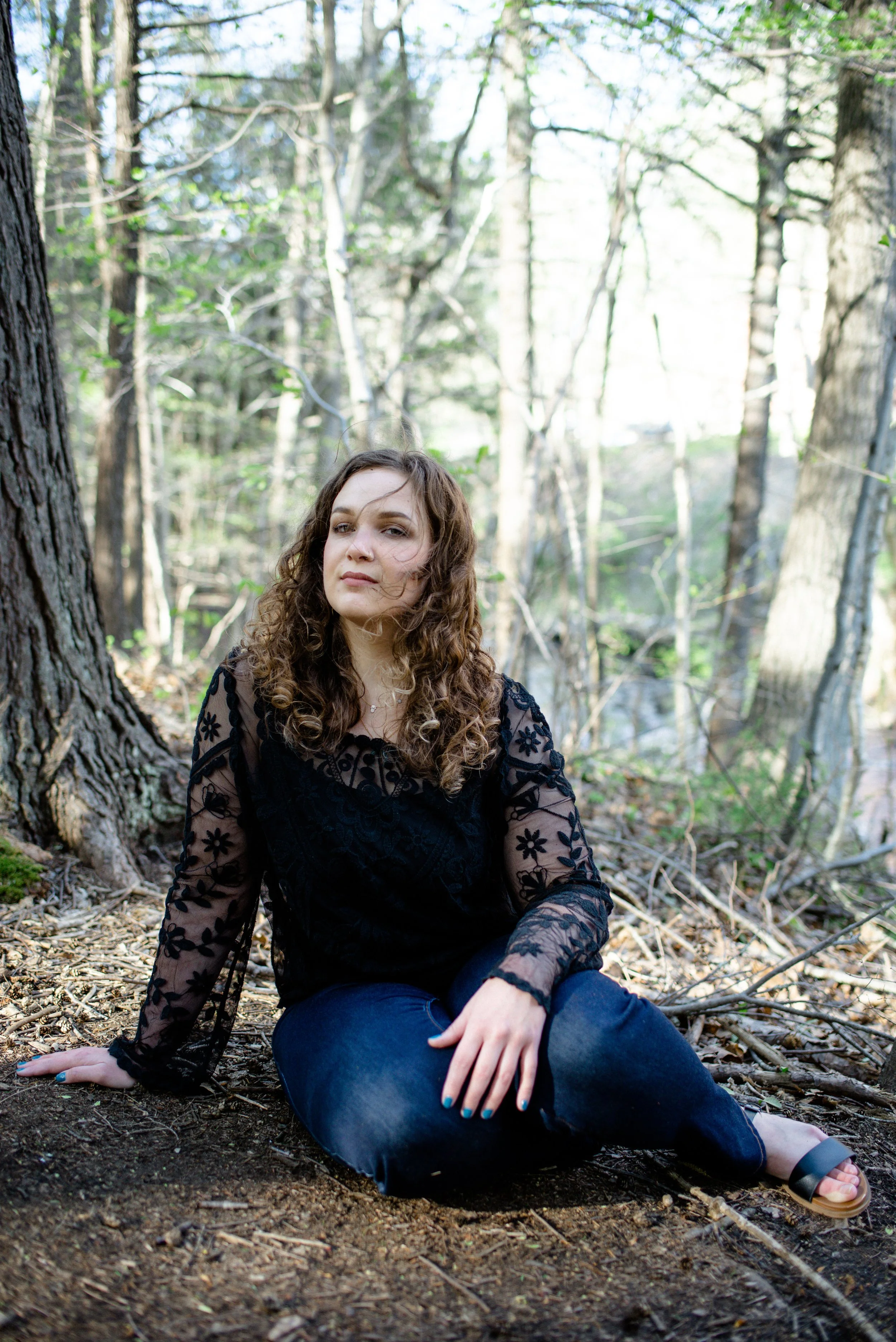 A woman with curly hair sitting on the ground in a forest, wearing a black lace top and blue jeans, with trees and sunlight in the background.