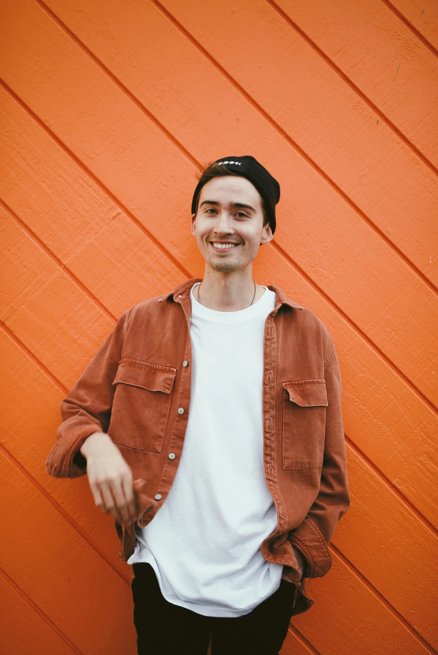 Smiling man in a black beanie and rust overshirt standing against an orange wooden wall
