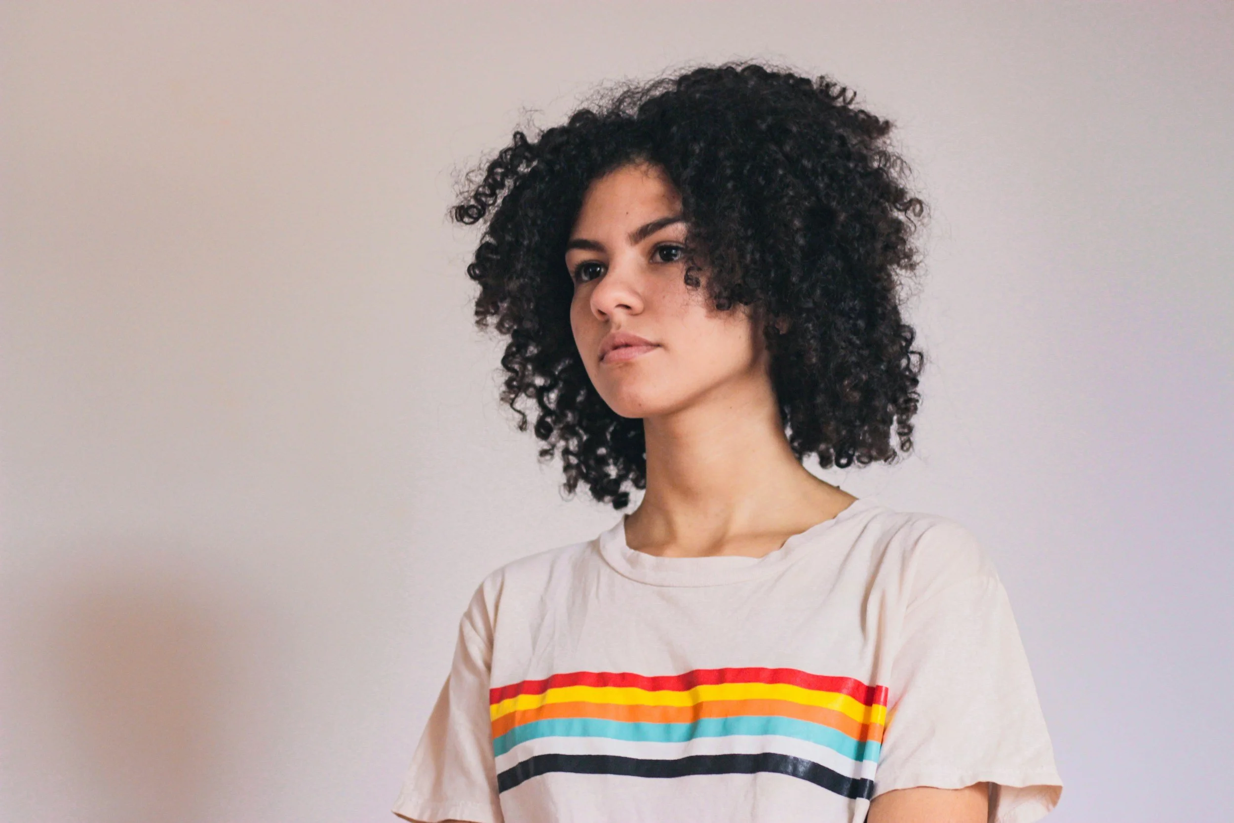 Young woman with curly black hair wearing a colorful striped T-shirt against a plain wall