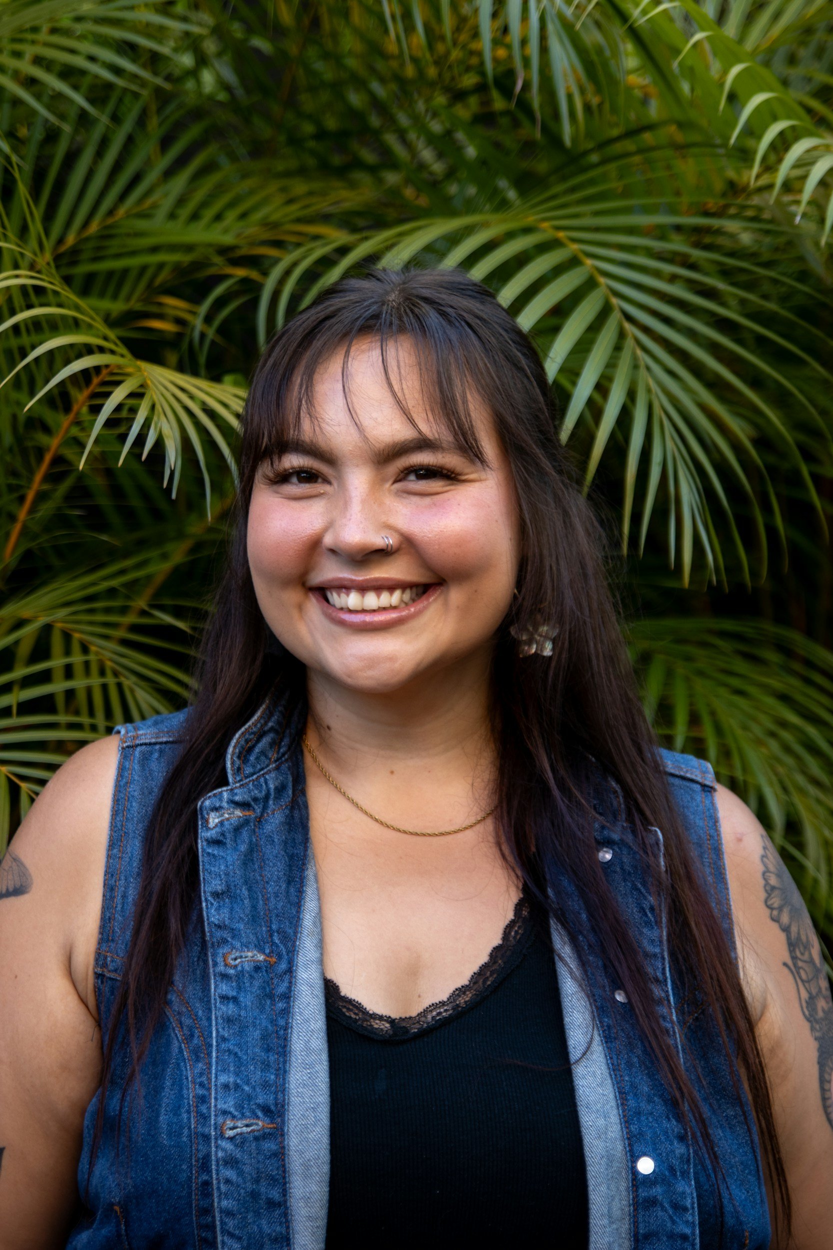 Smiling woman with long dark hair wearing a denim vest, standing in front of tropical palm leaves