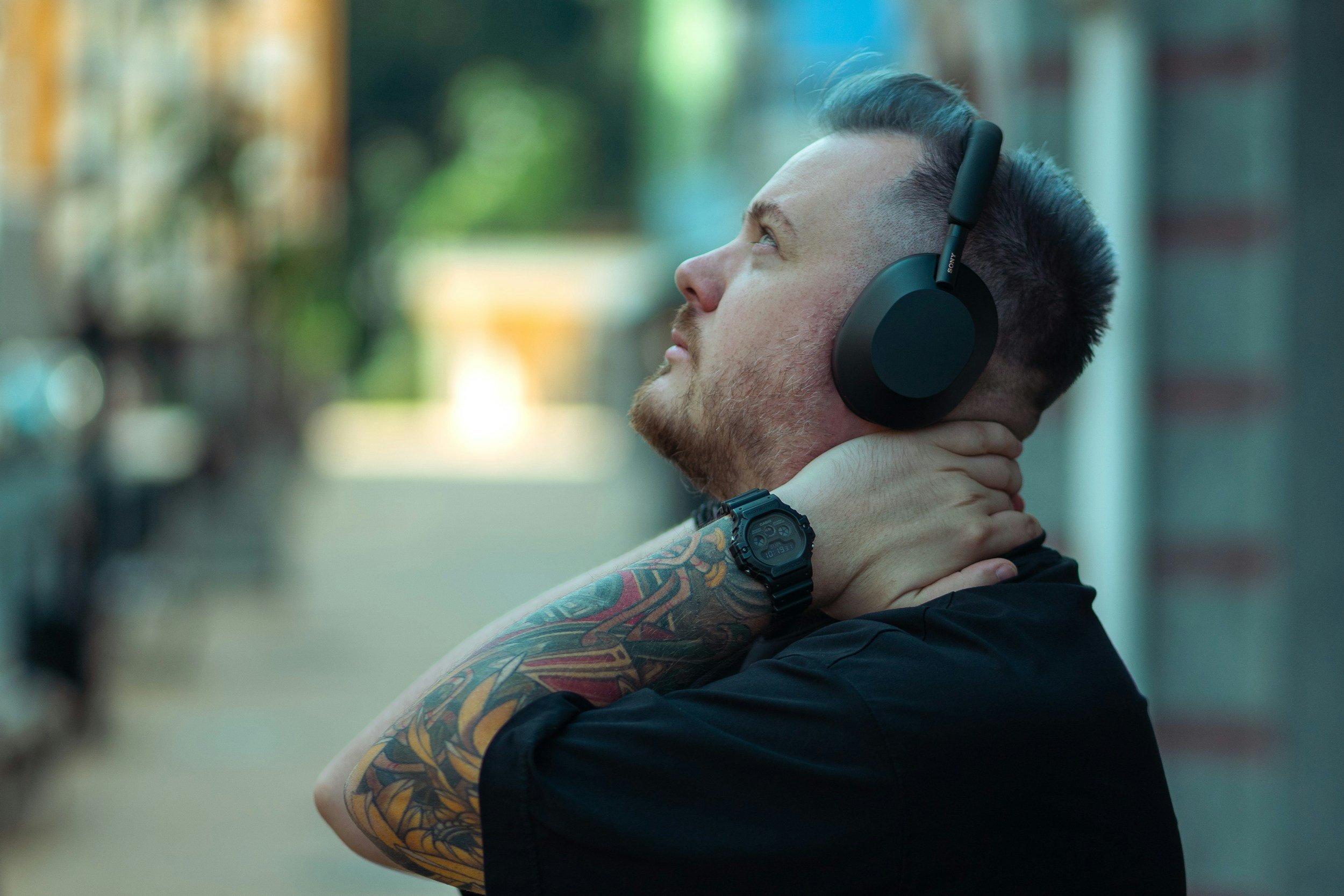 Tattooed man wearing black Sony headphones and a digital watch, viewed in profile against a blurred urban background