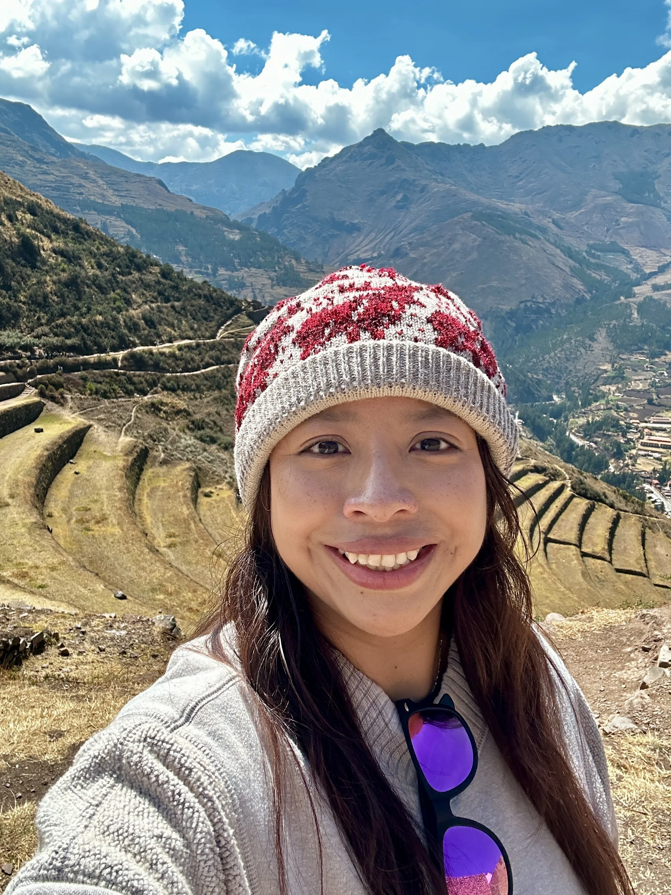 Smiling woman in a red-and-white knit hat taking a selfie with snow-capped mountains and terraced farmland behind her