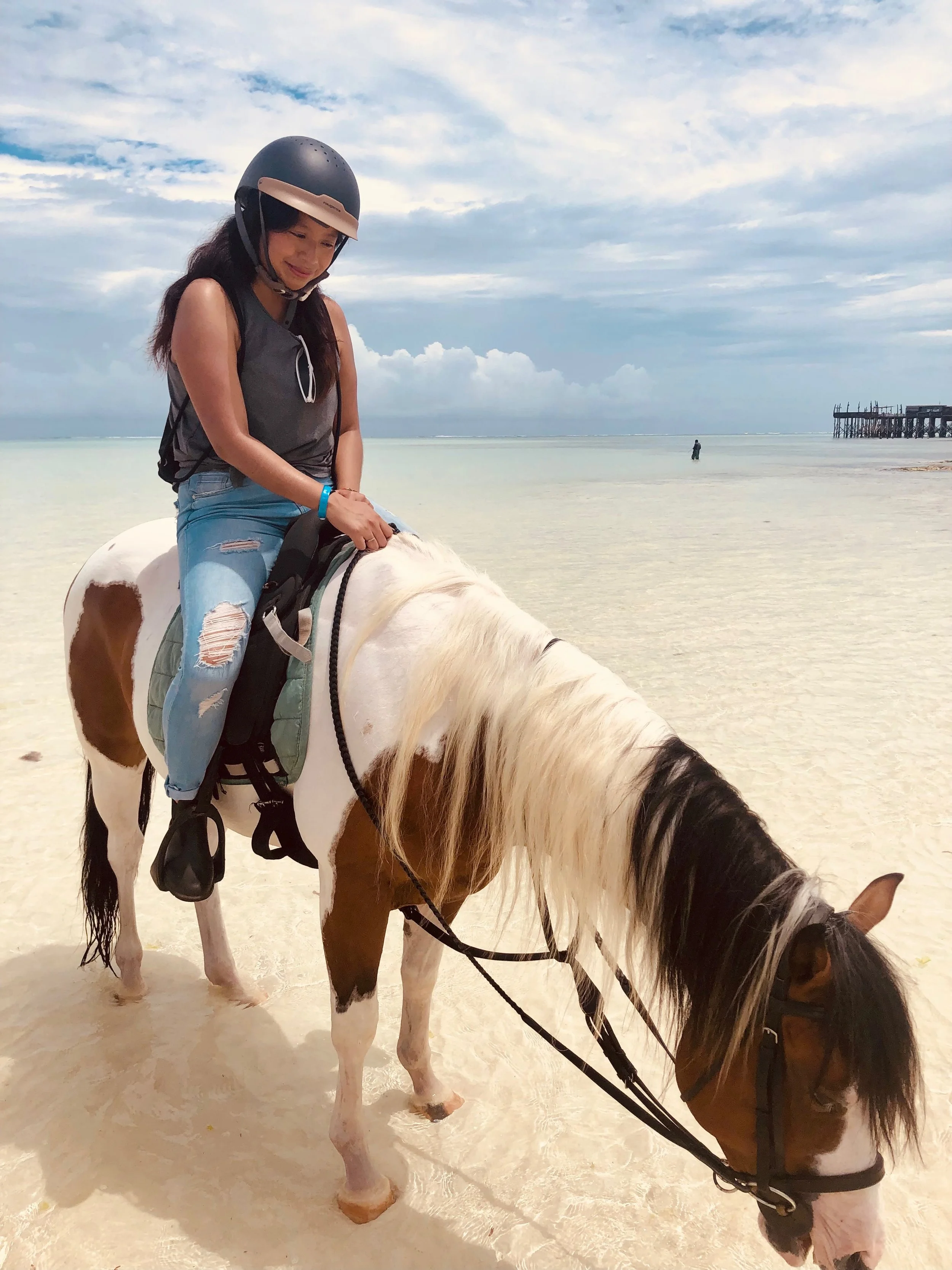 Woman wearing a helmet rides a white horse through shallow tropical beach water.