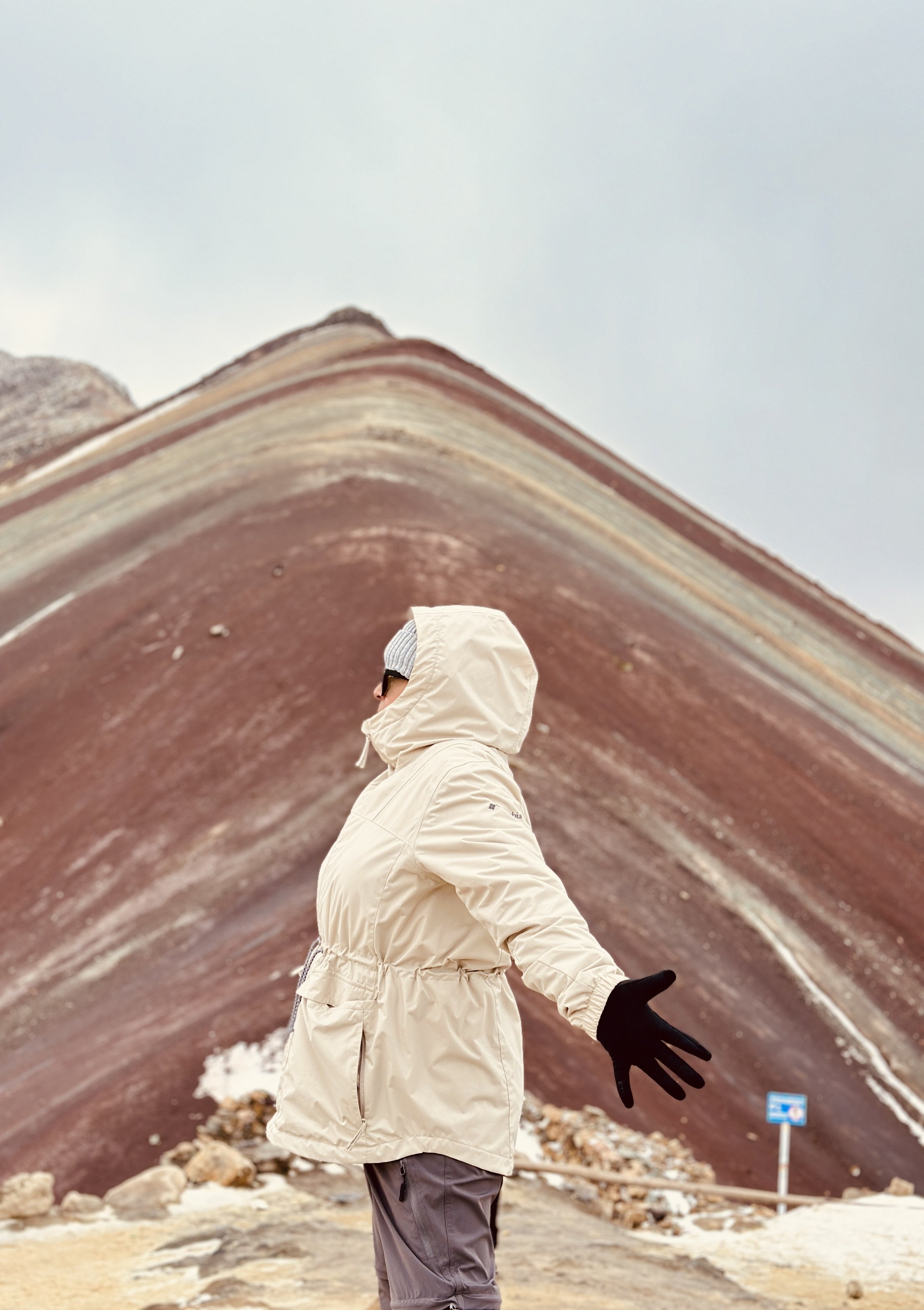Person in a white hooded winter jacket and black gloves standing before a reddish-brown volcanic crater.