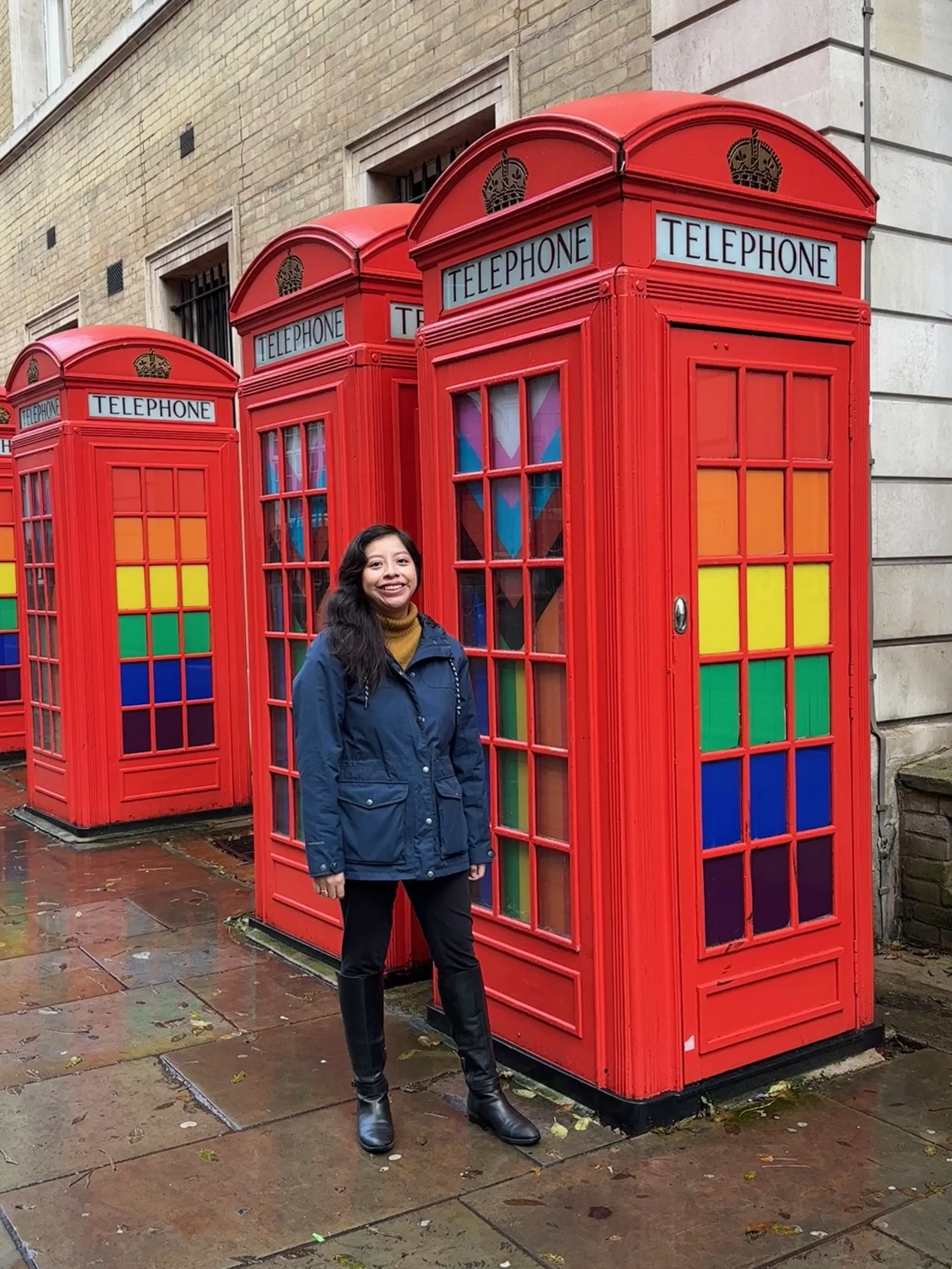 Woman posing in front of red telephone booths with the pride flag peaking through the windows.