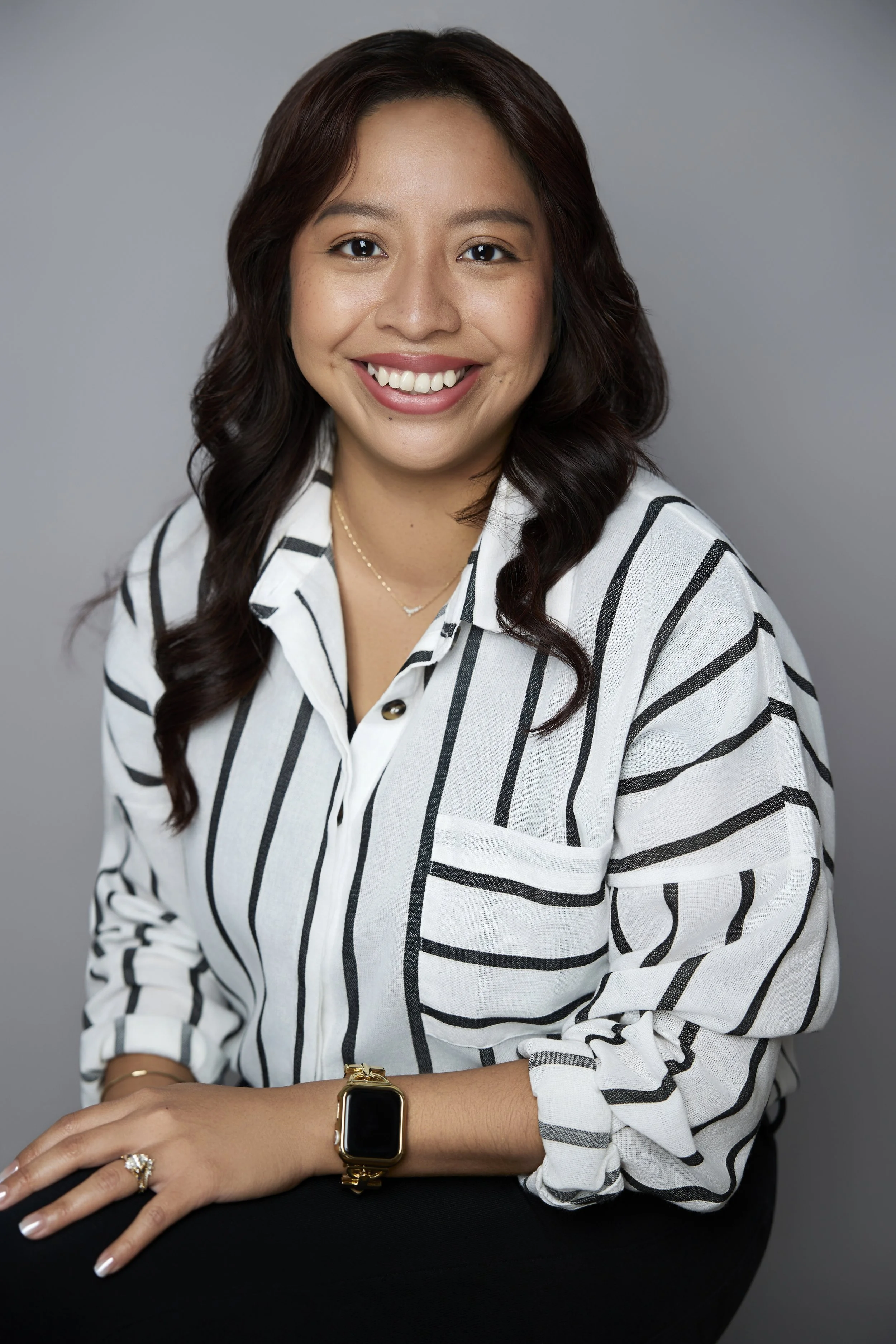 Smiling woman with long dark hair wearing a white and black patterned blouse, gold smartwatch, jewelry, and a ring against a gray background
