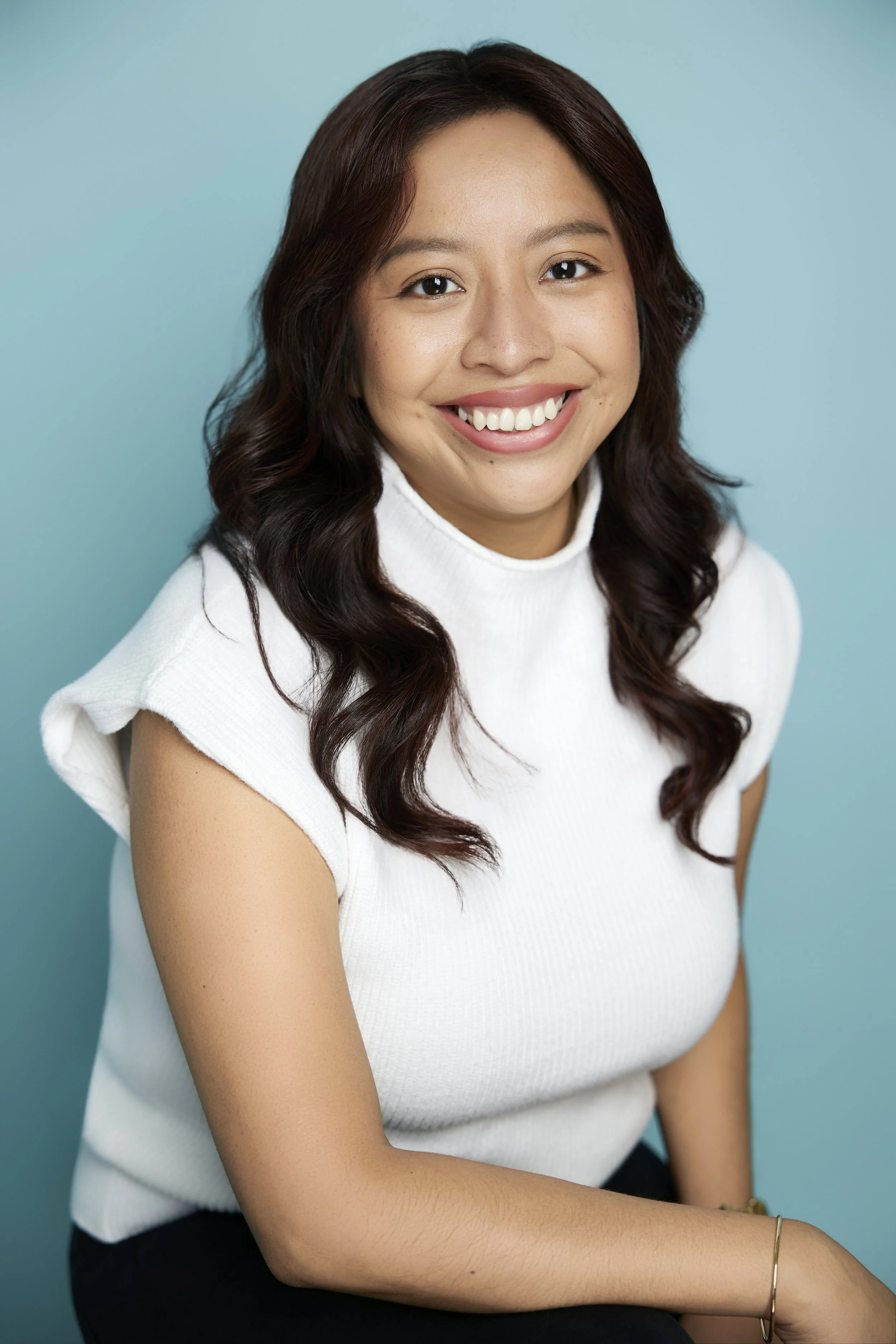 Smiling woman with long dark hair wearing a white shirt against a blue background