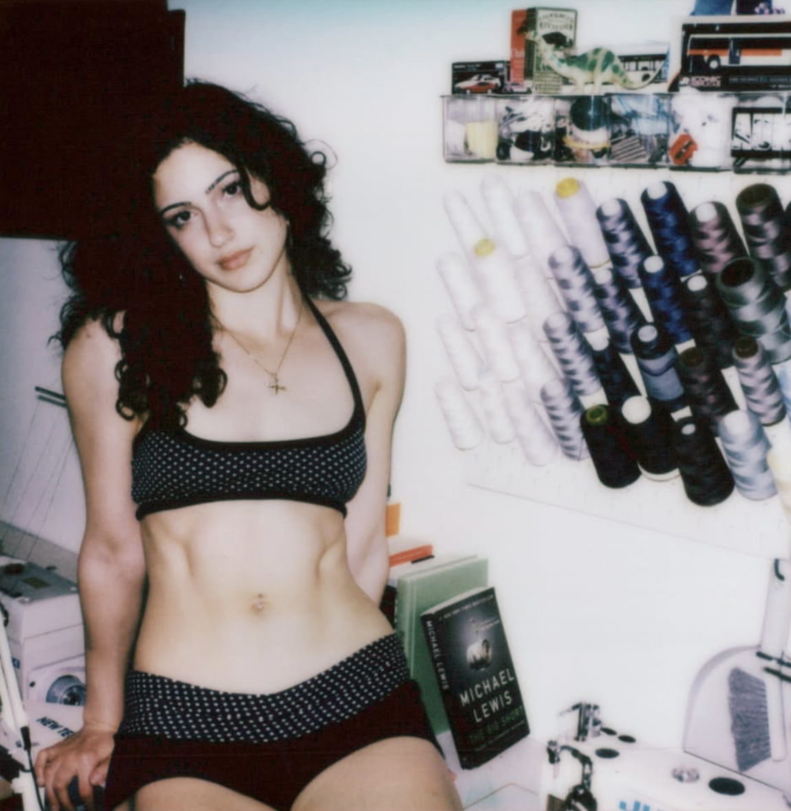 A young woman with curly dark hair and light skin standing in a room with shelves of spools of thread, a book on a table, and a small dinosaur toy on a shelf in the background.