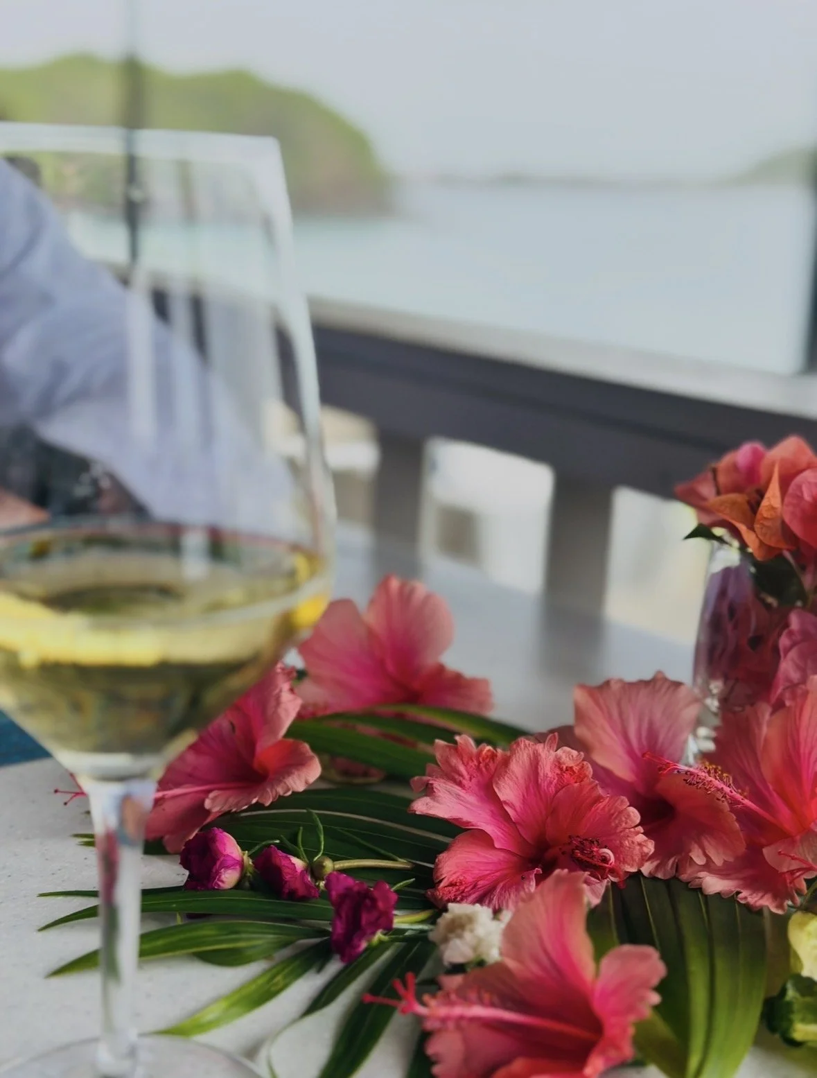 A close-up of a glass of white wine, pink hibiscus flowers, and a vase with additional pink flowers on a table with scenic ocean and greenery in the background.