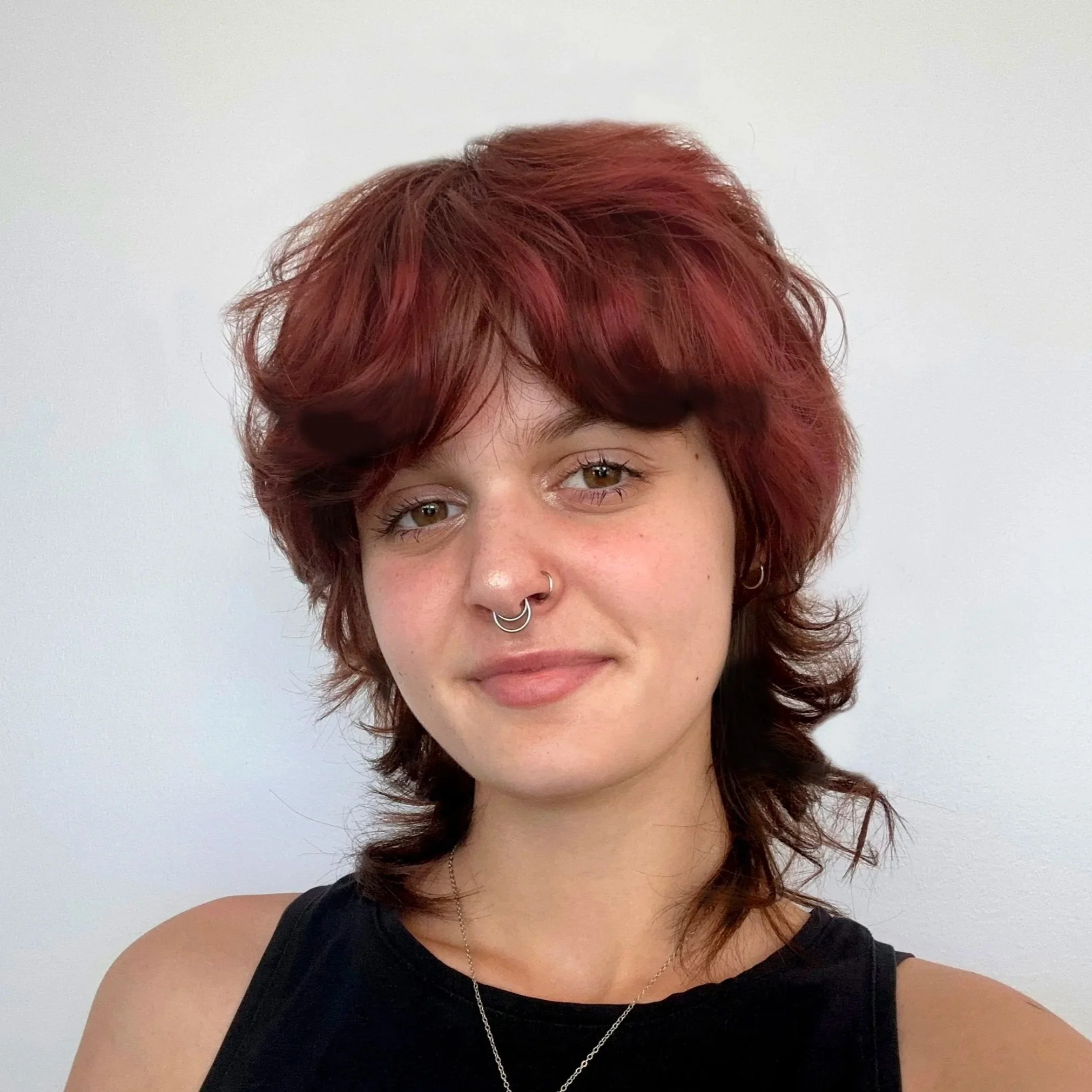 Close-up of a woman with short, wavy red hair, brown eyes, wearing a black sleeveless top, and a necklace, with a plain light-colored background.