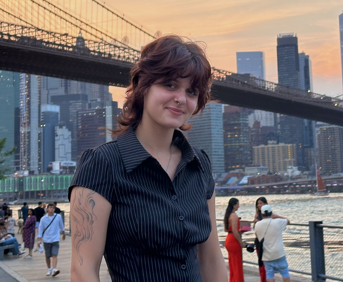 A young woman with short, wavy brown hair and a nose piercing stands outdoors near the water, with a city skyline and a suspension bridge in the background during sunset.