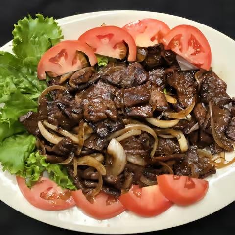 A plate of cooked dark meat with onions, surrounded by sliced tomatoes and green lettuce leaves.