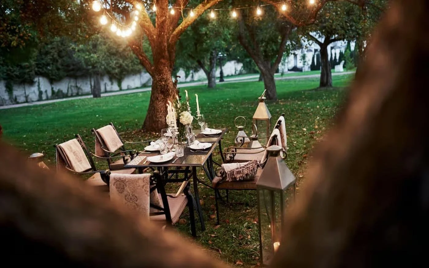 An outdoor dining setup with a table and chairs, decorated with lanterns and candles, situated under trees with string lights hanging, in a backyard or garden setting at dusk or evening.