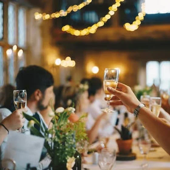 People raising glasses of champagne or wine in a toast at a dinner party with string lights and flowers in a warmly decorated room.