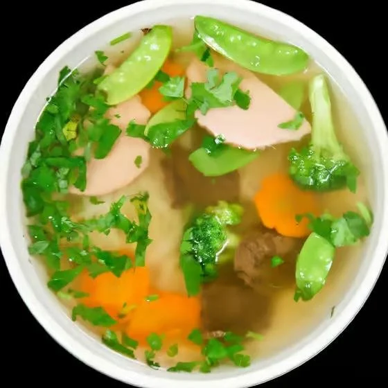 A bowl of hot, clear soup with slices of chicken, snow peas, carrots, beef, and chopped green herbs.