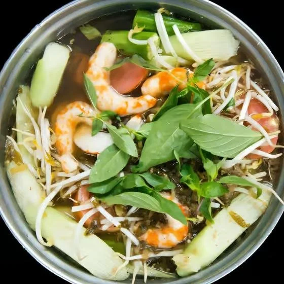 A bowl of Vietnamese pho with shrimp, basil, bean sprouts, green onions, and cucumber in broth.