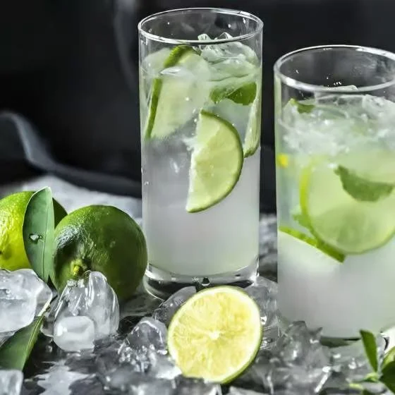 Two glasses of lime and cucumber infused water with ice, a halved lime, and fresh limes with ice cubes on a surface.