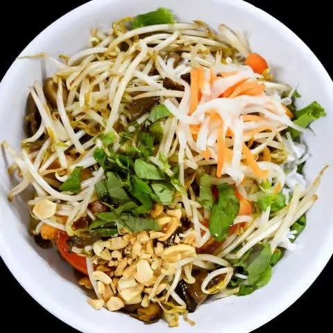 A bowl of noodle salad with bean sprouts, shredded carrots, chopped peanuts, and herbs.
