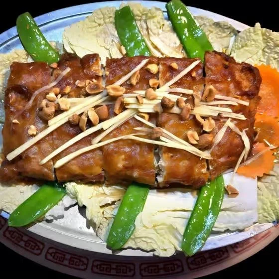 A plate of Asian-style fried fish covered in brown sauce, topped with chopped peanuts and shredded vegetables. Surrounding the fish are sliced green chili peppers and assorted sliced vegetables, including Napa cabbage and carrots.