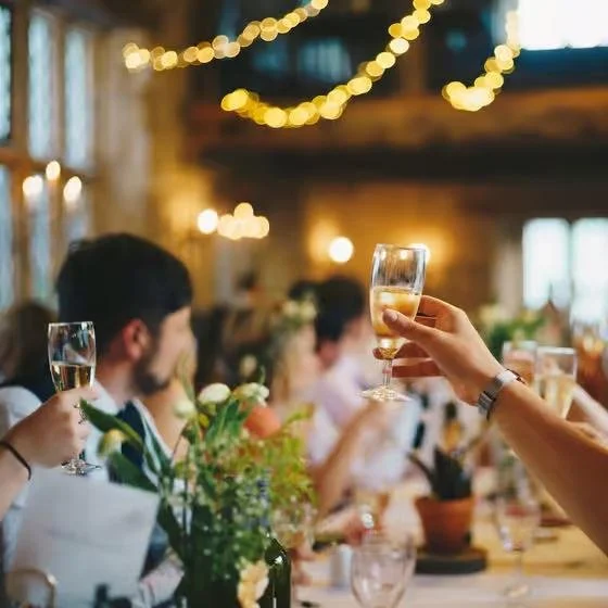 People raising glasses of champagne at a festive gathering in a warmly decorated room with string lights and flowers.