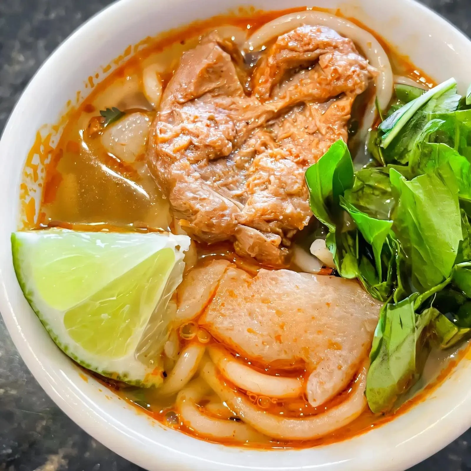 A bowl of Vietnamese Pho noodle soup with slices of meat, fresh herbs, lime wedges, and rice noodles.