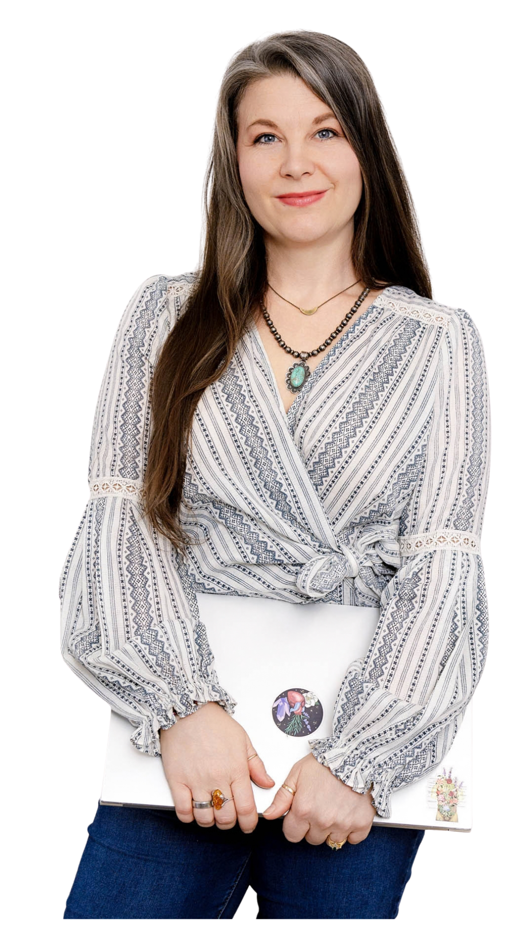 A woman with long brown hair wearing a patterned wrap blouse and jeans, holding a white laptop decorated with stickers, standing against a plain background.