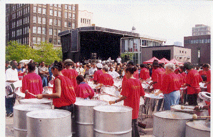 A crowd of people wearing red shirts playing steel drums at an outdoor event in a city park.
