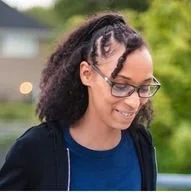 A woman with glasses and natural curly hair smiling outdoors