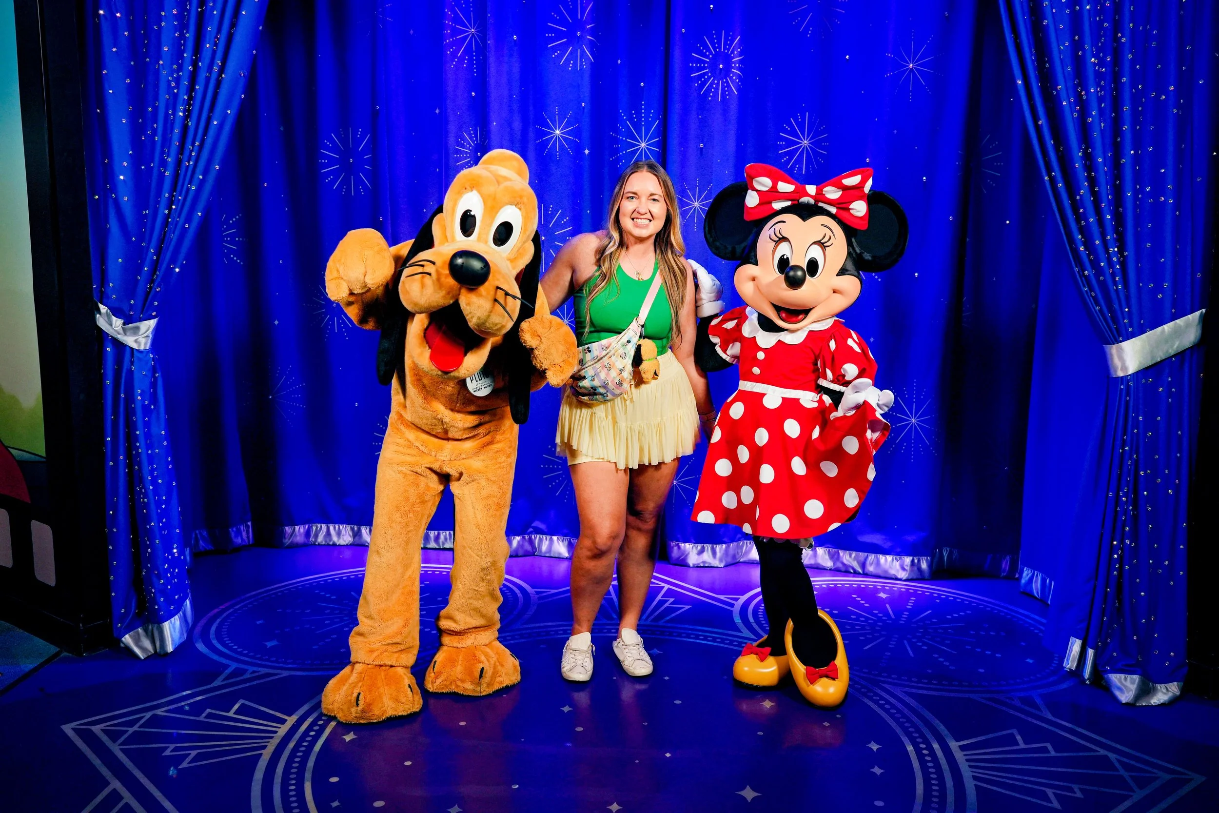 A woman standing between life-size costumes of Pluto and Minnie Mouse, posing in front of a blue curtain with starburst patterns.