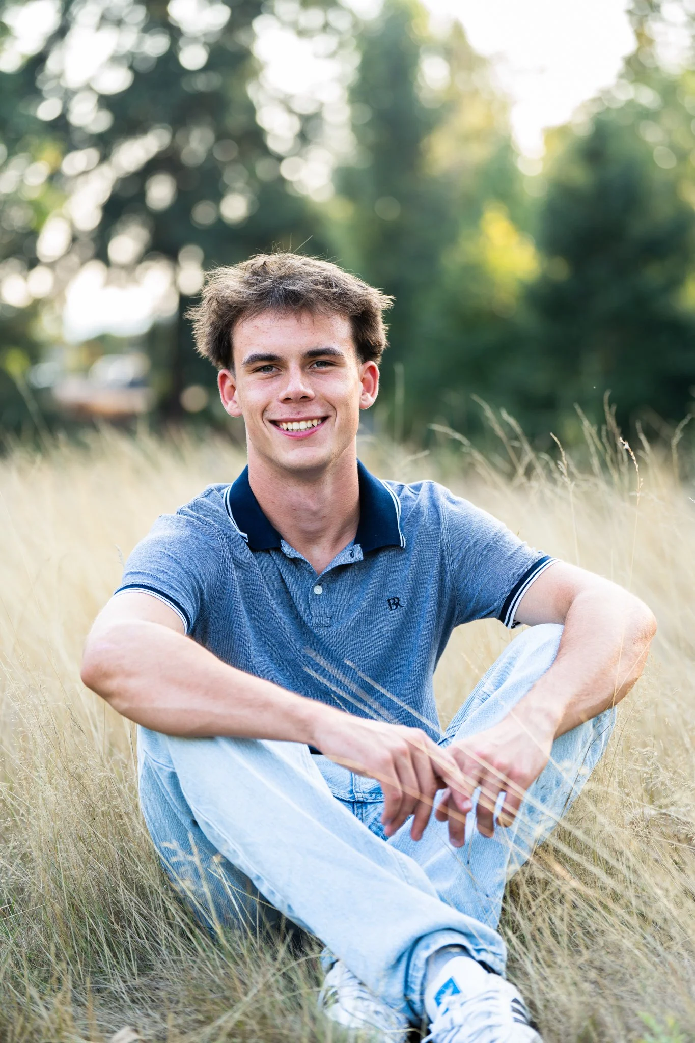 A young man with brown hair smiling, sitting in a field of tall grass with trees in the background during daytime.